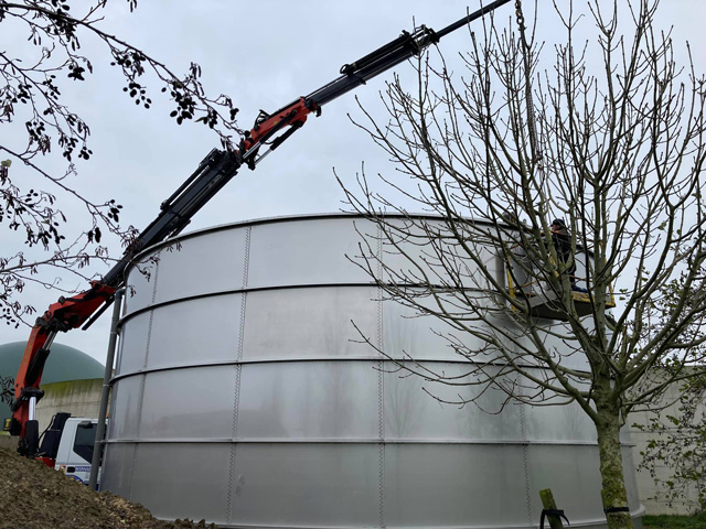Zwart-oranje vrachtwagenkraan met knikarm hijst een grote stalen tank op een terrein