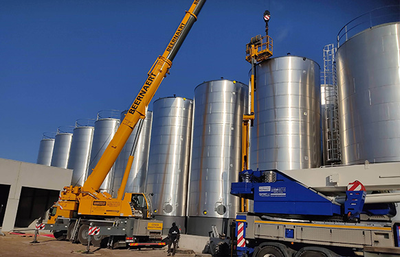 Gele telescopische Beernaert-kraan met uitgeschoven giek bij grote roestvrijstalen silo's